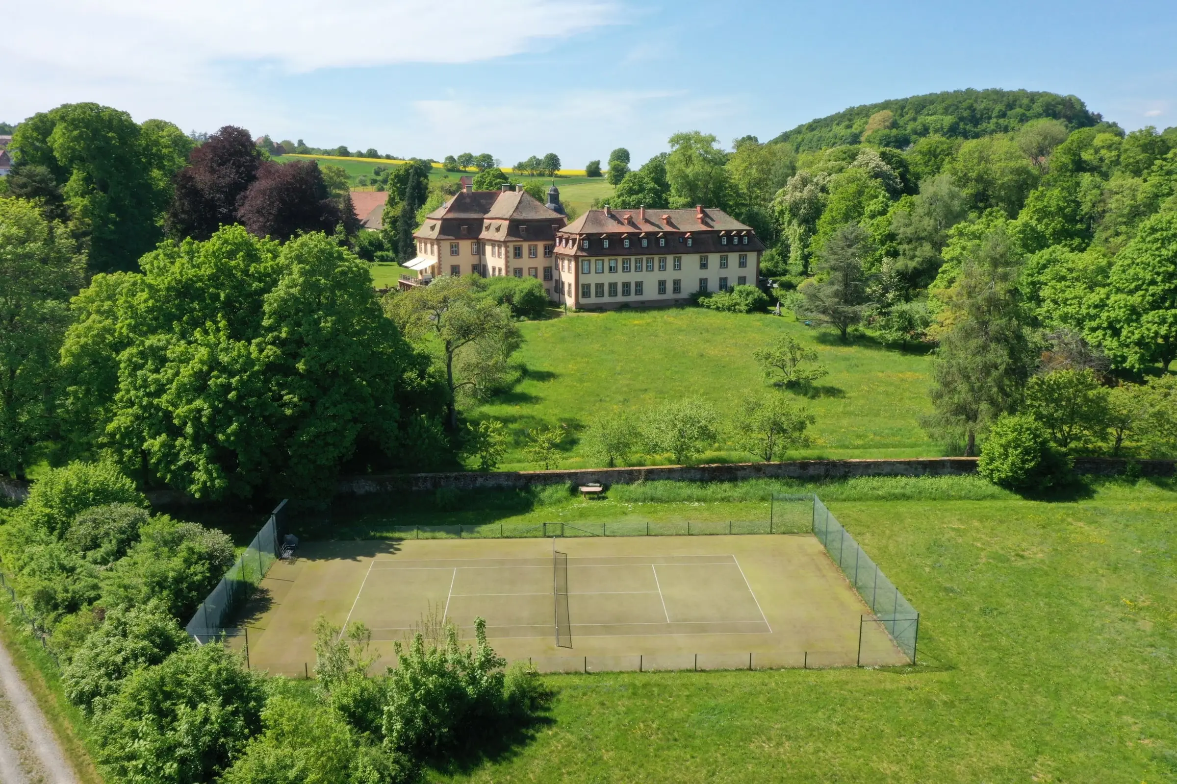 Luftaufnahme von Schloss Roßbach am Waldrand, umgeben von Wiesen, im Vordergrund der Tennisplatz